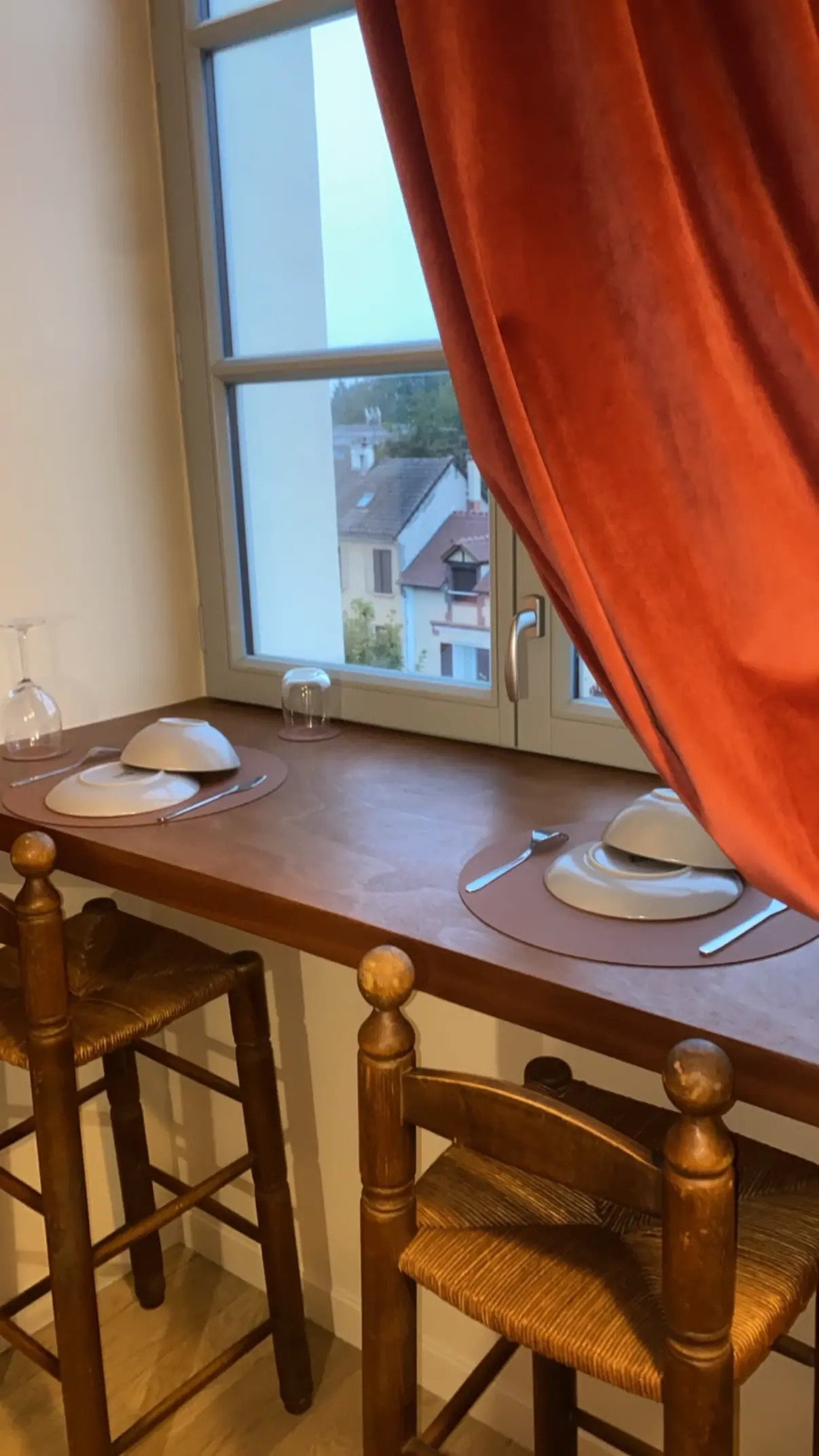 Comptoir en bois devant une fenêtre, avec assiettes et verres dressés pour deux et un rideau orange